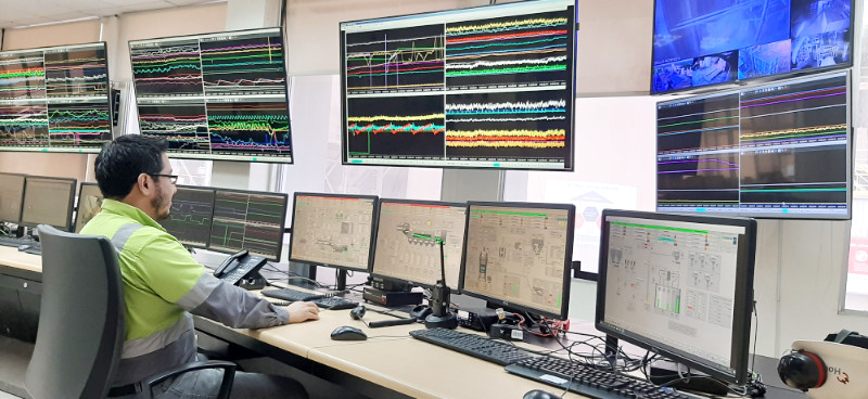 A person in a high-visibility jacket monitors multiple screens displaying data and graphs in a control room.